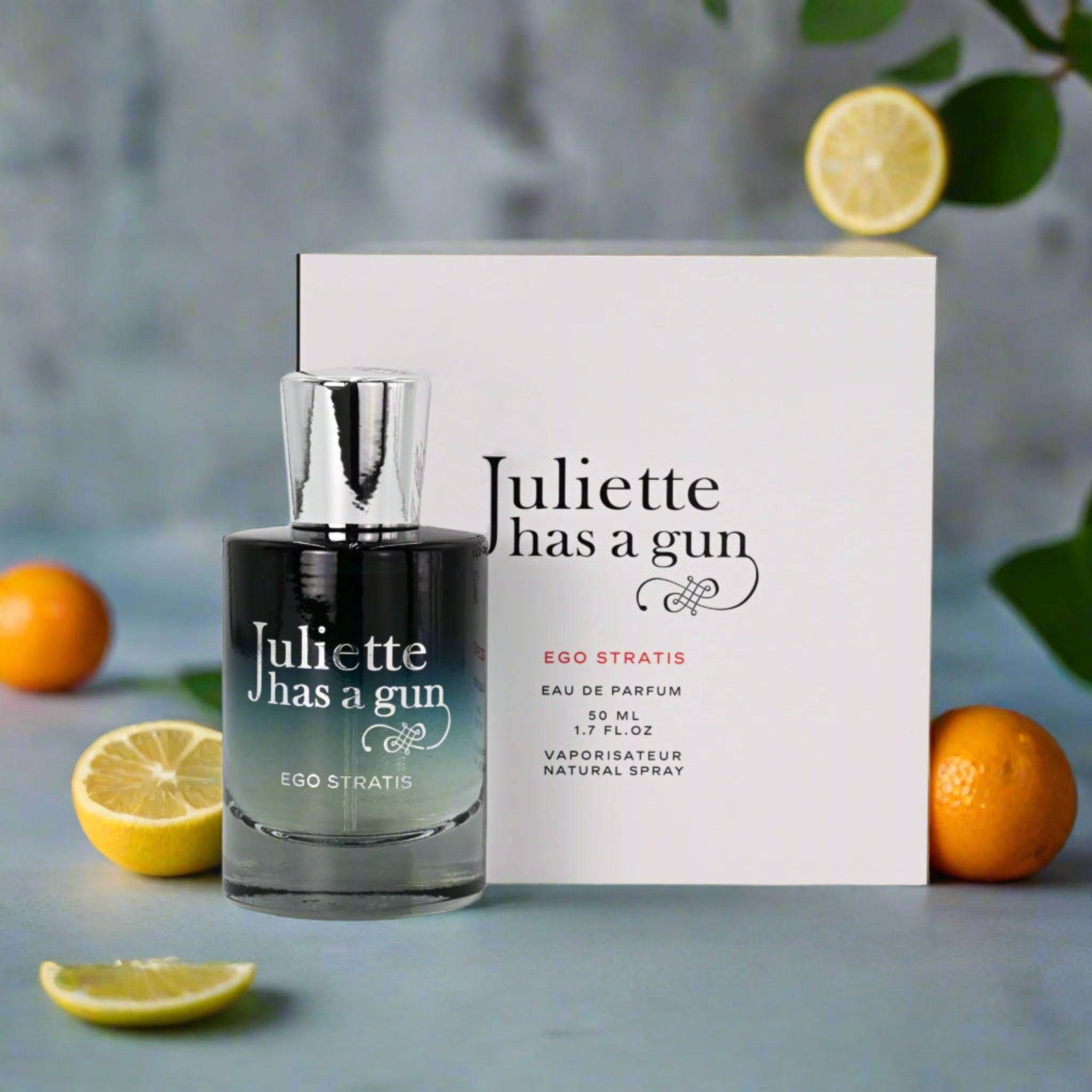 Perfume bottle and packaging of 'Juliette has a gun' with lemons and oranges on a gray background
