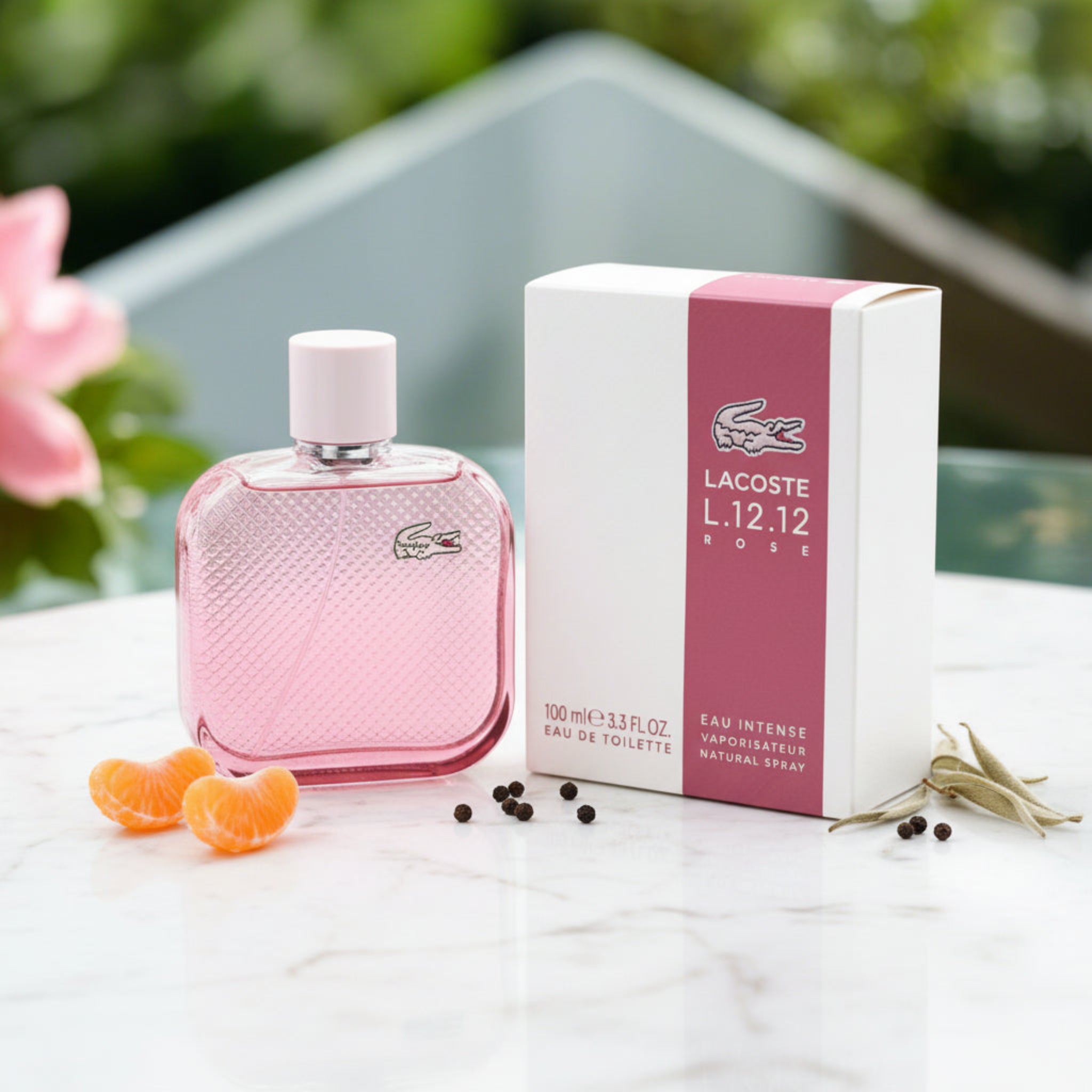 Pink Lacoste L.12.12 Rose perfume bottle and packaging on a reflective surface with blurred greenery in the background.