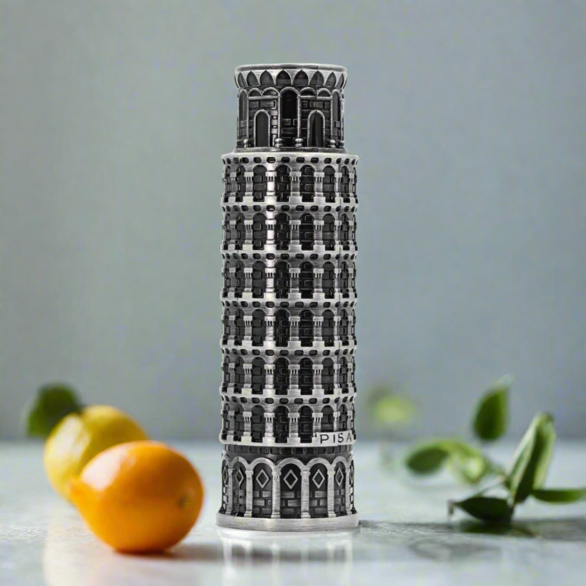 Perfume bottle designed like the Leaning Tower of Pisa on a table with fruits and leaves.