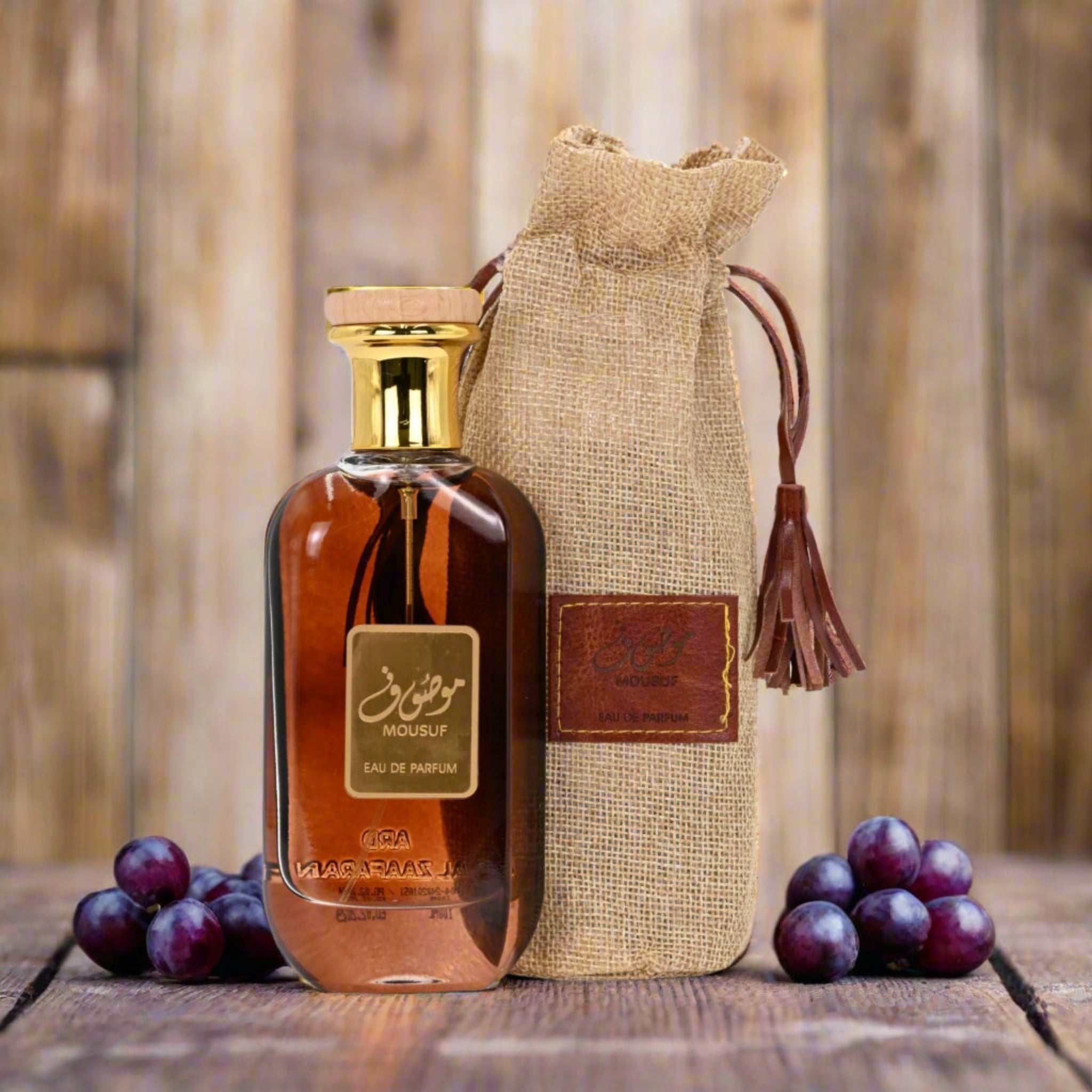 Brown perfume bottle with gold cap and decorative hessian bag on a wooden surface with grapes.