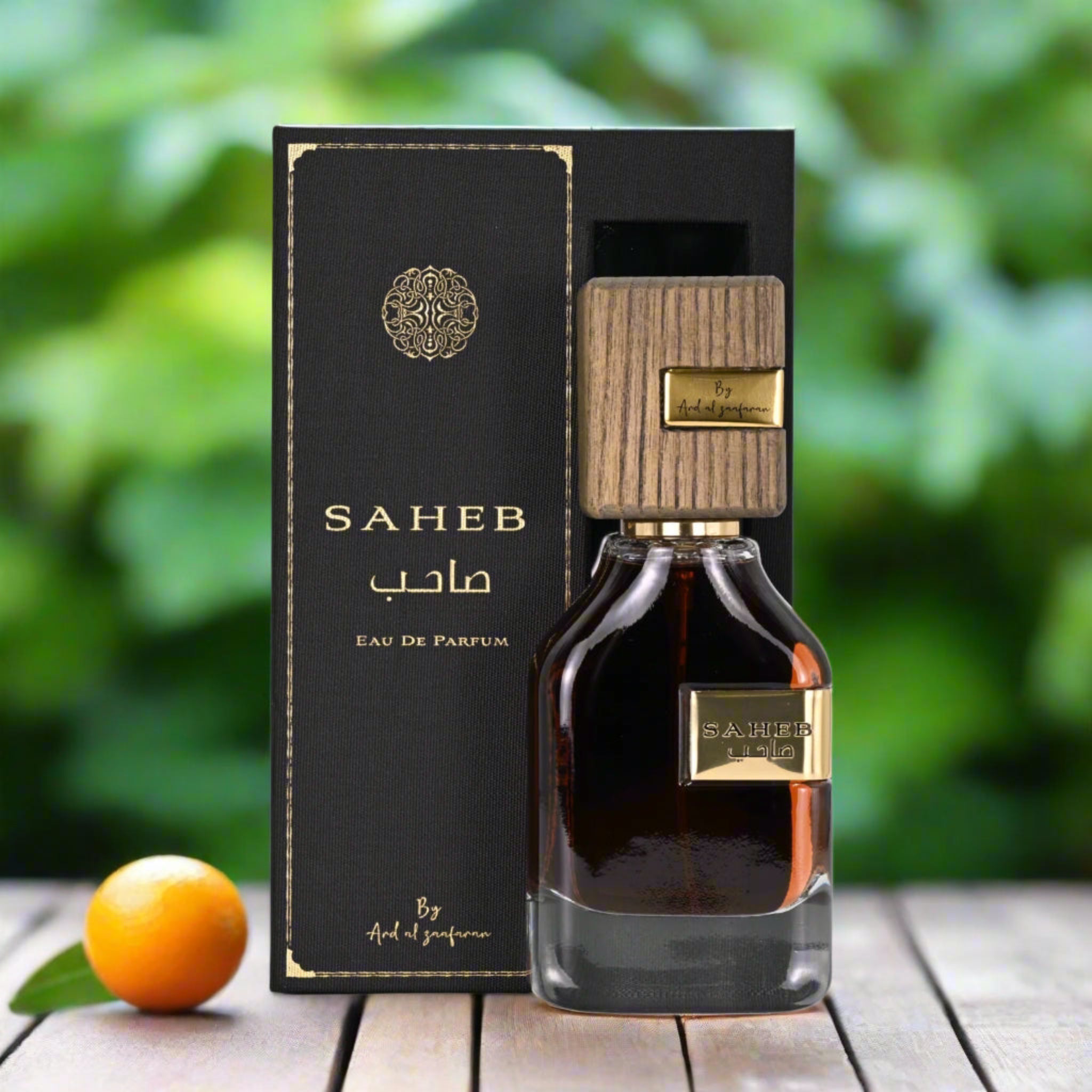 SAHEB Eau De Parfum bottle and packaging on a wooden surface with a blurred green background