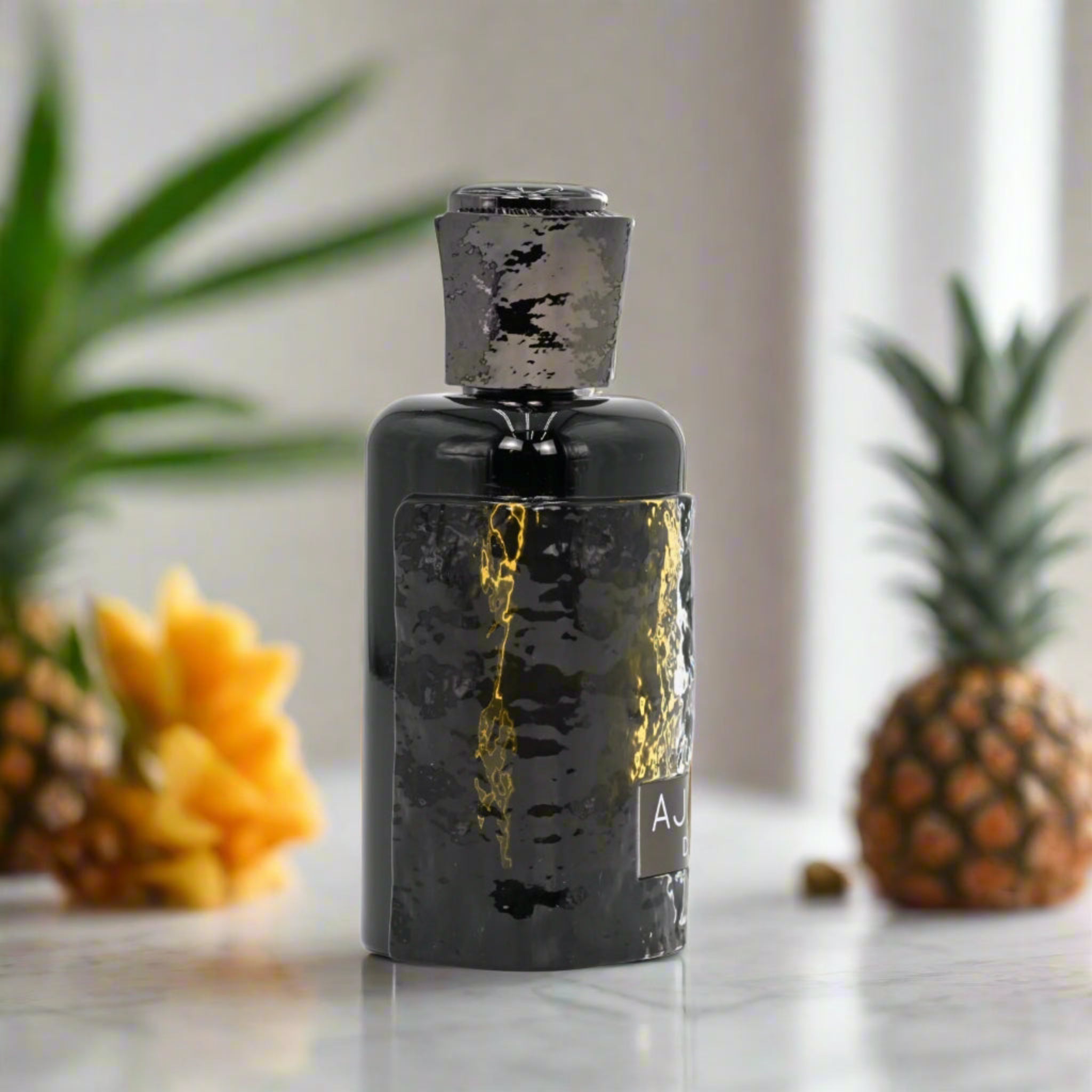 Black bottle with gold accents on a marble surface with pineapples and flowers in the background