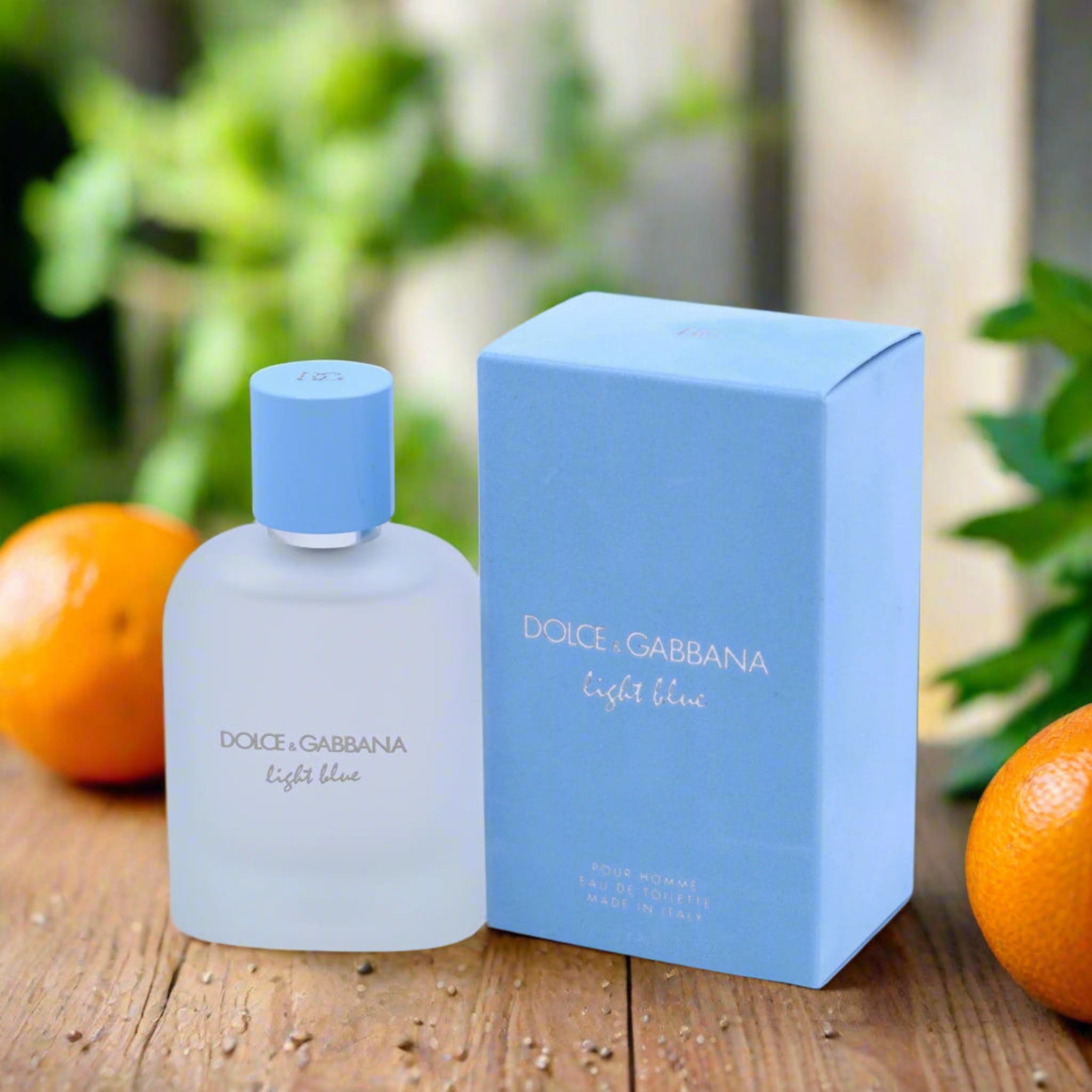 Dolce & Gabbana Light Blue perfume bottle and packaging on a wooden surface with oranges and greenery in the background.