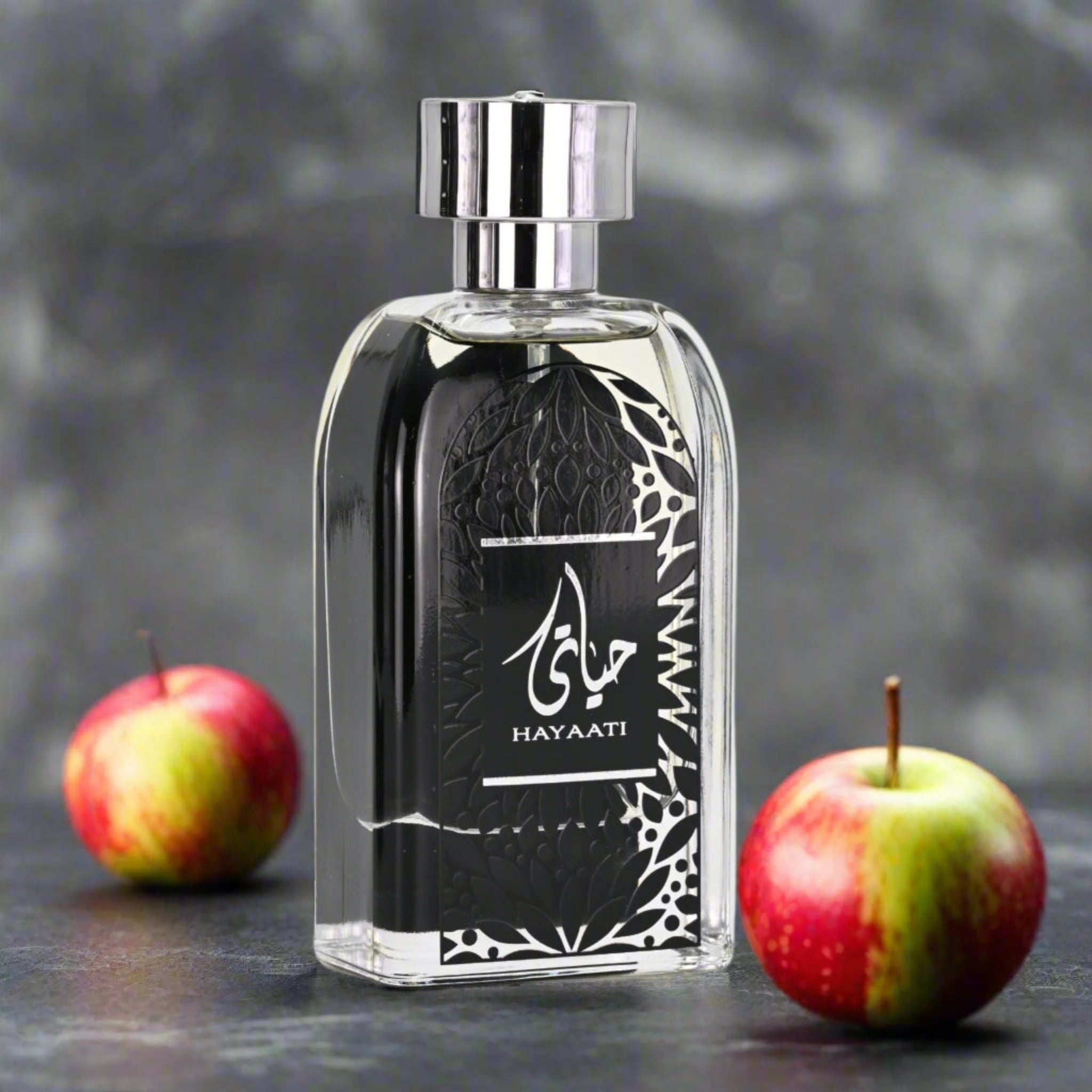 Perfume bottle with 'Hayaati' branding next to two apples on a dark surface.