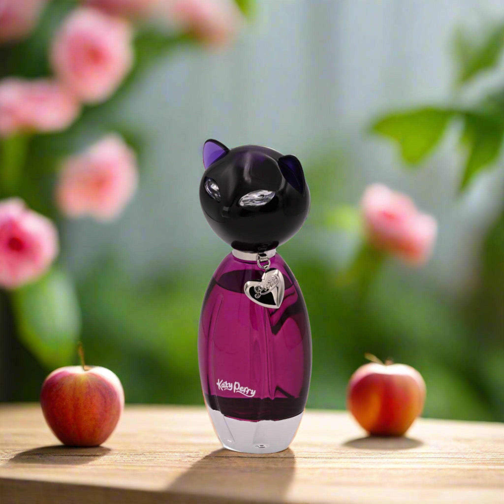 Cat-shaped purple perfume bottle with apples on a wooden surface and blurred flowers in the background