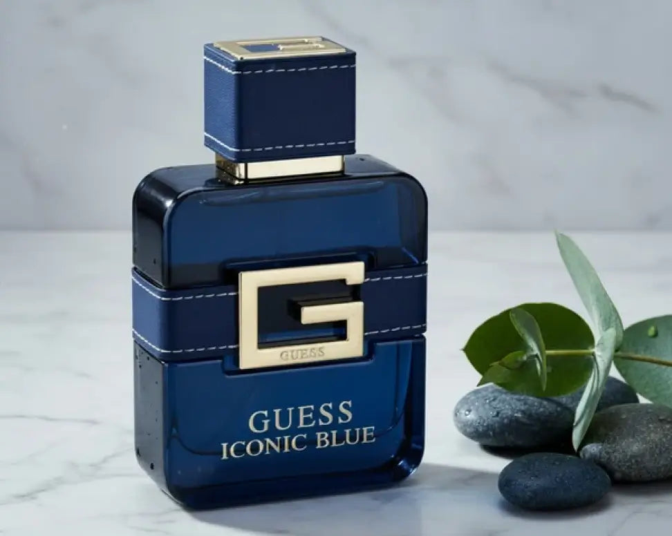 Guess Iconic Blue men’s cologne in a navy bottle with a gold G buckle, beside gray stones and eucalyptus leaves.