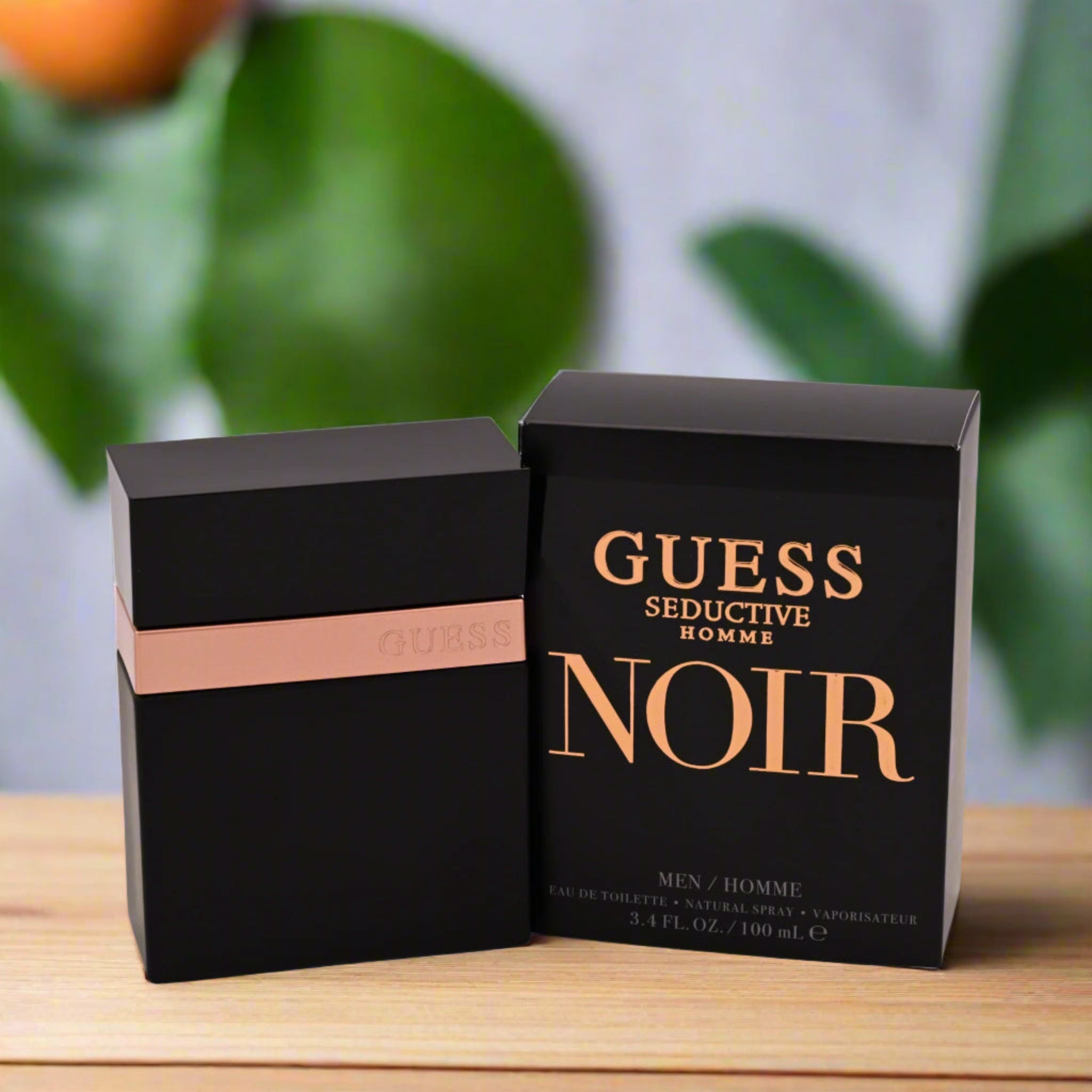 Guess Seductive Homme Noir perfume packaging on a wooden surface with a blurred green plant background