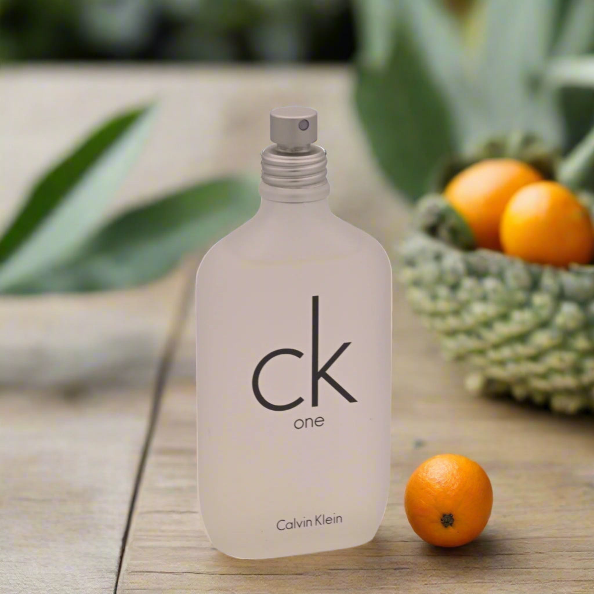Calvin Klein CK One perfume bottle on a wooden surface with fruits and plants in the background