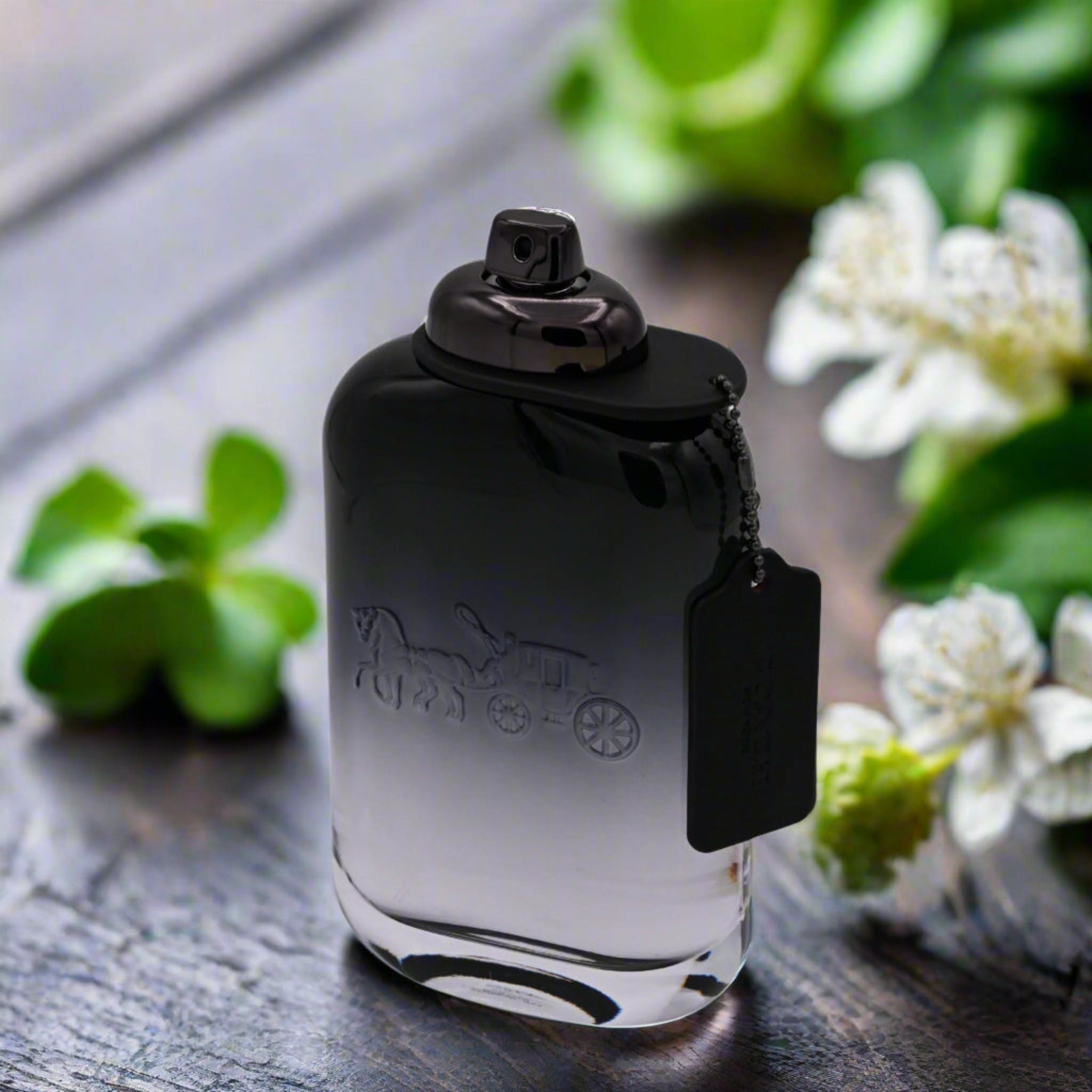 Black perfume bottle with a brand logo on a wooden surface with greenery and flowers in the background