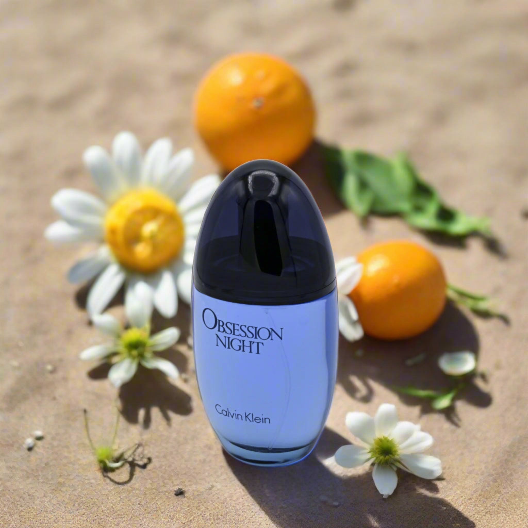 Calvin Klein Obsession Night perfume bottle on sand with flowers and oranges