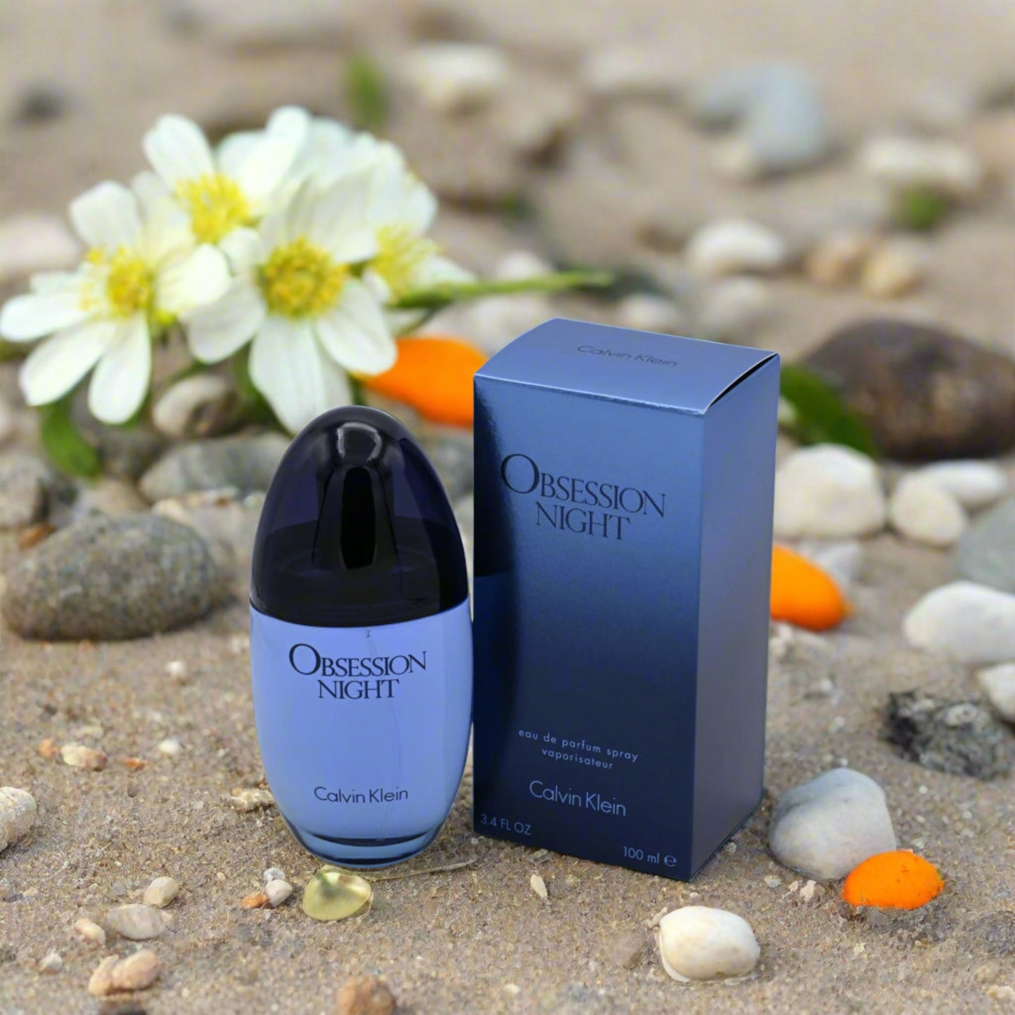 Calvin Klein Obsession Night perfume bottle and packaging on a sandy background with flowers and pebbles.